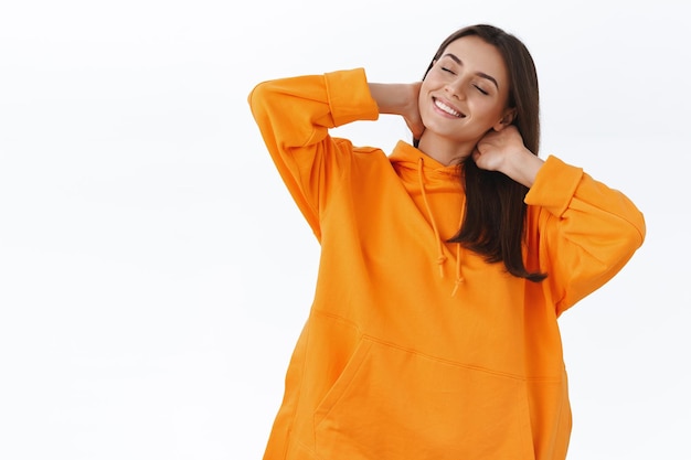 Vrije tijd, weekends en mensen concept. ontspannen zorgeloos jong mooi meisje in oranje hoodie, haar nek aanraken met gesloten ogen en tevreden glimlach