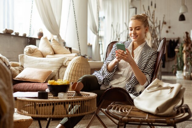 Vrijetijdszone. Stralend blonde vrouw rusten in comfortabele ruime fauteuil en chatten op een smartphone