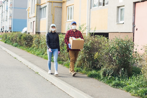 Vrijwilligers in beschermende maskers lopen over straat met een doos boodschappen, liefdadigheid.