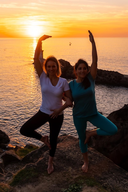 Vrksasana twee vrouwen die meditatie en yoga doen op een rots bij zonsondergang naast een vuurtoren in de zee, gezond en naturistisch leven