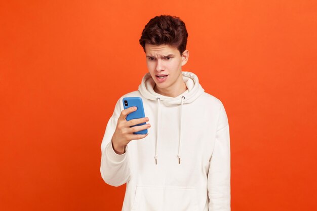 Vroeg zich af tiener in wit casual sweatshirt met capuchon kijkend naar smartphonescherm met verbaasd gezicht, slechte verbinding. indoor studio-opname geïsoleerd op oranje achtergrond