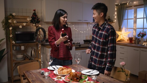 Vrolijk Aziatisch echtpaar dat bij de feestelijke eettafel staat en de kerstwijn bespreekt die ze aan de vooravond van de vakantieviering zullen hebben