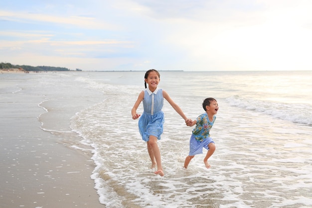 Foto vrolijk aziatisch kindmeisje en kleine jongen die plezier hebben, rennen samen en hand in handen op tropisch zandstrand bij zonsondergang gelukkige familie geniet van de zomervakantie