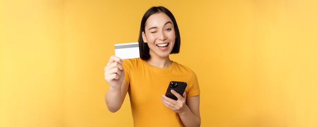 Vrolijk aziatisch meisje dat lacht met creditcard en smartphone die mobiel bankieren aanbeveelt tegen een gele achtergrond
