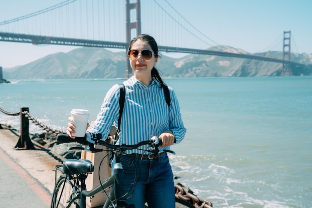 vrolijk Aziatisch meisje met fiets en wandelen met koffie geniet van het landschap in de jachthaven met golden gate bridge en blauwe zee op de achtergrond in Californië.