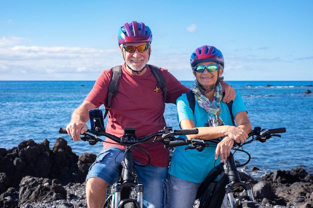 Vrolijk blank senior paar poseren voor een portret op het strand rijden op elektrische fietsen Authentiek pensioenleven en gezonde levensstijl concept Horizon over de zee
