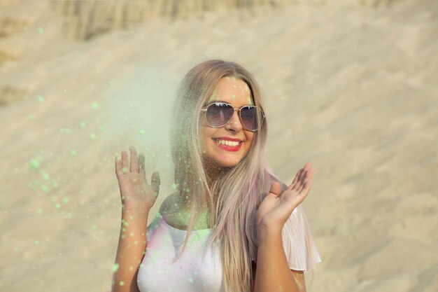 Vrolijk blond model met een bril die poseert met exploderende groene Holi-verf in de woestijn