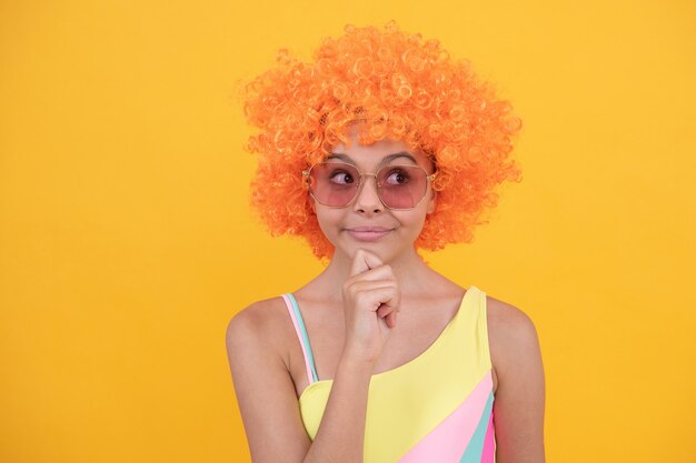 Vrolijk denkend tienermeisje in zonnebril en zwempak met oranje krullend pruikhaar op gele achtergrond, emoties.