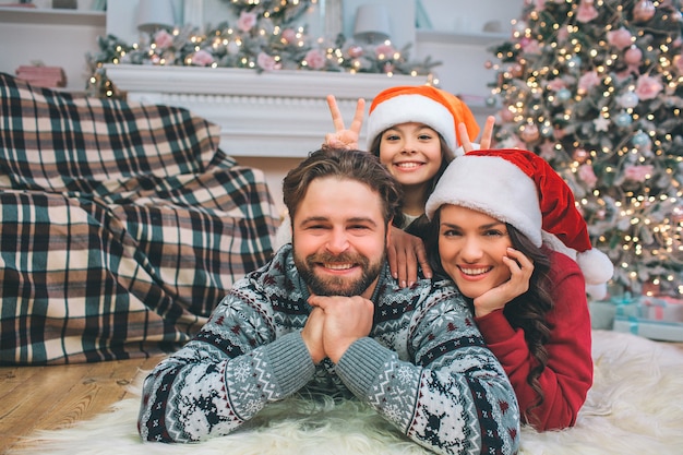 Vrolijk en positief jong gezin dat op de vloer ligt en er recht uit ziet. Ze lachen. Jonge vrouw en meisje staan achter de man. Ze dragen kerstmutsen.