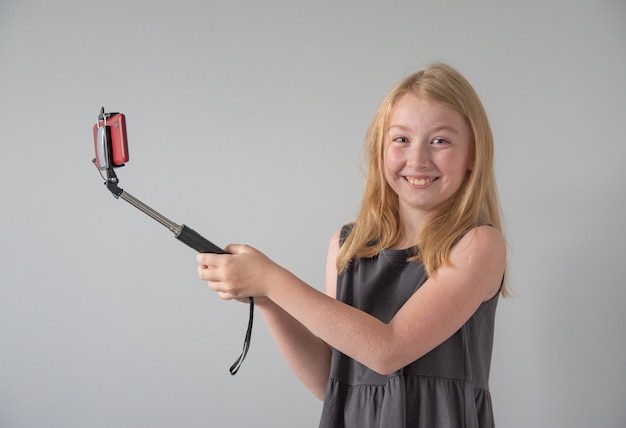 Vrolijk en vrolijk meisje in een grijze jurk met een selfiestick en een rode telefoon maakt een grappige foto of video op een grijze achtergrond