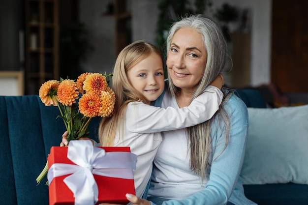 Vrolijk europees klein meisje dat oude vrouw omhelst met geschenkdoos en boeket bloemen in het interieur van de woonkamer