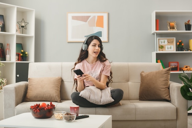 Vrolijk geknipperd jong meisje met een koptelefoon met een telefoon op de bank achter de salontafel in de woonkamer