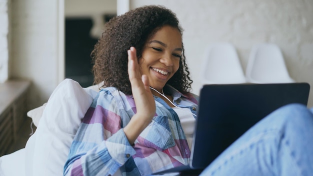 Vrolijk gemengd rasmeisje dat videochat met vrienden die een laptopcamera gebruiken terwijl ze op bed ligt