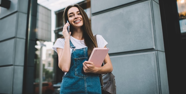 vrolijk hipstermeisje in casual jeanskleding die bij camera glimlacht tijdens vriendschappelijke smartphone