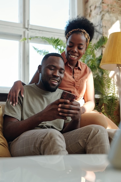 Vrolijk jong Afrikaans stel in vrijetijdskleding die met een glimlach naar het smartphonescherm kijkt terwijl ze voor de camera tegen een groot raam zitten