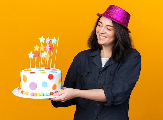 Vrolijk jong kaukasisch feestmeisje met een feesthoed die cake uitrekt met sterren die ernaar kijken, geïsoleerd op een oranje muur