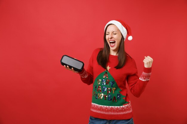 Vrolijk jong kerstmanmeisje dat winnaargebaar doet, met draagbare draadloze bluetooth-muziekluidspreker geïsoleerd op rode achtergrond. Gelukkig Nieuwjaar 2019 viering vakantie partij concept. Bespotten kopie ruimte.