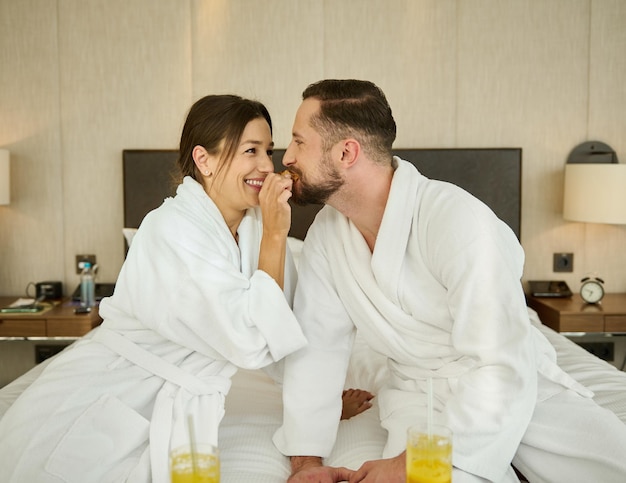 Vrolijk jong liefdevol getrouwd stel, jonggehuwden zittend op het bed na het wakker worden en genieten van een heerlijk ontbijt voor twee in een slaapkamer van een luxe hotel terwijl ze hun huwelijksverjaardag vieren