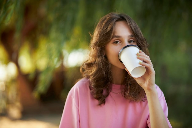 Vrolijk jong meisje in een roze t-shirt loopt in het park en drinkt afhaalkoffie uit een papieren bekertje.