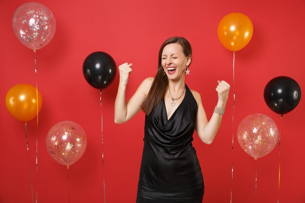 Vrolijk jong meisje in kleine zwarte jurk vieren, winnaar gebaar doen op heldere rode achtergrond luchtballonnen. St. Valentijnsdag, Vrouwendag, Gelukkig Nieuwjaar, verjaardag mockup vakantie feest concept.