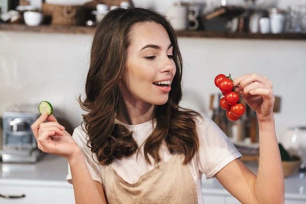 Vrolijk jong meisje met een schort dat thuis in de keuken zit en een tros tomaten vasthoudt