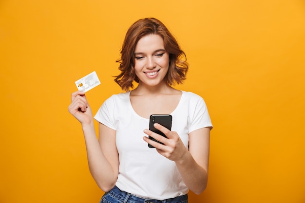 Vrolijk jong meisje met een t-shirt dat geïsoleerd over een gele muur staat, met behulp van mobiele telefoon, met plastic creditcard