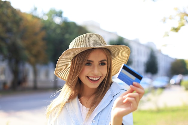 Vrolijk jong optimistisch meisje dat buiten staat en een creditcard in de hand houdt.