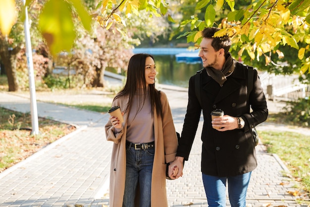 Vrolijk jong stel dat in de herfst leuke tijd doorbrengt in het park, hand in hand loopt, afhaalkoffie drinkt