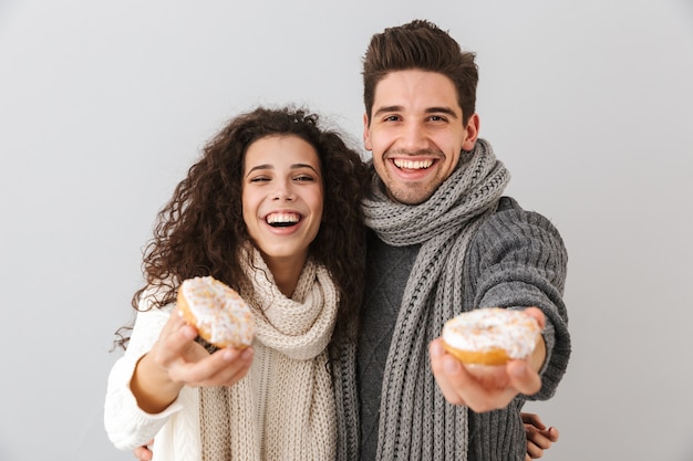 Vrolijk jong stel dat truien en sjaals draagt die zich geïsoleerd over grijze muur bevinden, die donuts tonen