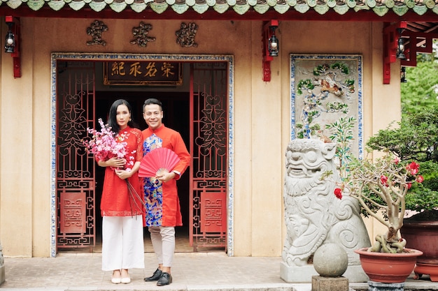 Vrolijk jong Vietnamees stel in traditionele jurken die bij tempeldeuren staan met bloeiende perziktakken en papieren waaier