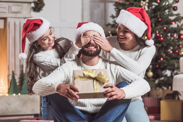 Vrolijk kerstfeest en een gelukkig nieuwjaar! Knappe man houdt een cadeautje vast en lacht terwijl zijn dochter en vrouw zijn ogen bedekken