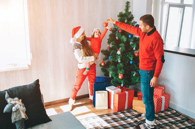 Vrolijk kerstfeest en een gelukkig nieuwjaar. Mooi en helder beeld van een jong gezin dat bij de kerstboom staat. De mens houdt rood stuk speelgoed en glimlacht. Kind reikt er met belangstelling naar uit.