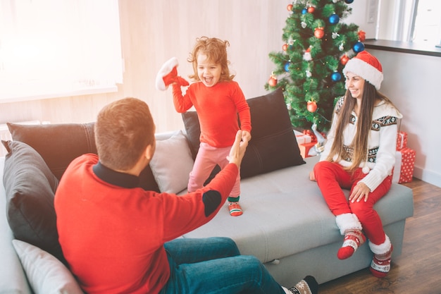 Vrolijk kerstfeest en een gelukkig nieuwjaar. Speelse foto van een gelukkig kind dat op de bank staat en rode hoed houdt. Ze kijkt vader aan en schreeuwt. Jonge vader houdt de hand van zijn dochter vast. .