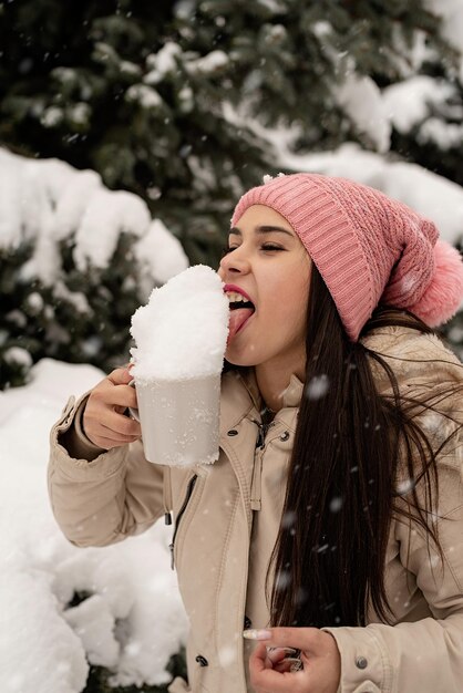 Vrolijk kerstfeest en een gelukkig nieuwjaar. winterplezier en vreugde. vrouw in warme winterkleren die buiten bij de grote kerstboom staat en sneeuw uit de beker likt