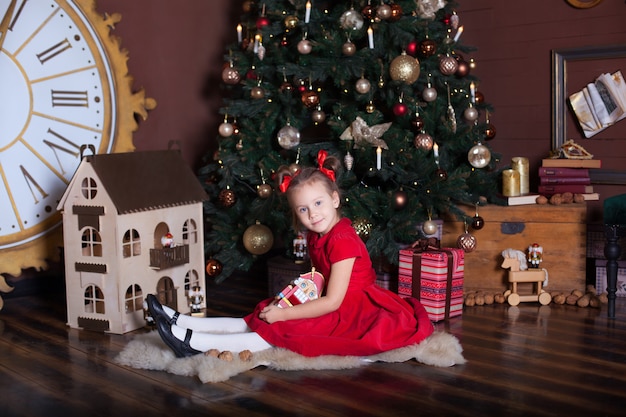 Vrolijk kerstfeest, fijne feestdagen. Klein meisje in een rode vintage jurk zit in de buurt van een versierde kerstboom met een houten speelgoed De notenkraker. Vakantie met het gezin. Gelukkig kind geniet van de vakantie.