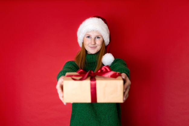 Vrolijk kerstfeest portret van een mooi jong tienermeisje in een gezellige gebreide groene trui en Santa39s hoed met geschenkdozen De rode achtergrond is de plaats voor de tekst