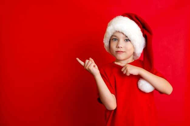 Vrolijk Kerstfeest. Portret van een schattige vrolijke jongen in een kerstmuts in een rood T-shirt op een rode achtergrond. Een plek voor tekst. Hoge kwaliteit foto