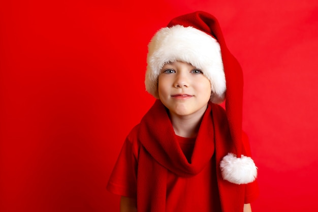 Vrolijk Kerstfeest. Portret van een schattige vrolijke jongen in een kerstmuts in een rood T-shirt op een rode achtergrond. Een plek voor tekst. Hoge kwaliteit foto