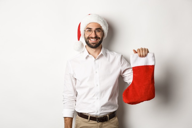 Vrolijk kerstfeest, vakantie concept. Gelukkig volwassen man ontvangt geschenken in xmas sok, opgewonden kijken, met kerstmuts, witte achtergrond.