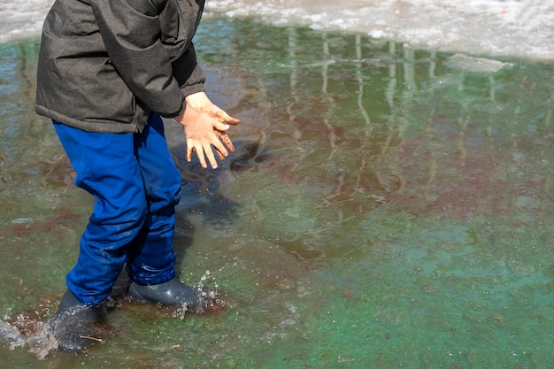 Vrolijk kind in rubberen laarzen vuil kneden met handen in modderige plas Milieuvervuiling Kind jongen van 67 jaar oud wandelen en spelen in laarzen in vervuild water