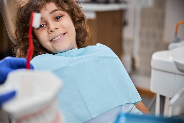 Vrolijk kind zit in tandartsstoel terwijl hij kijkt naar een demonstratie tandenpoetsen door zijn tandarts by