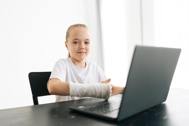 Vrolijk klein meisje met gebroken hand gewikkeld in wit gipsverband met laptopcomputer die thumbup laat zien terwijl ze naar de camera zit aan het bureau
