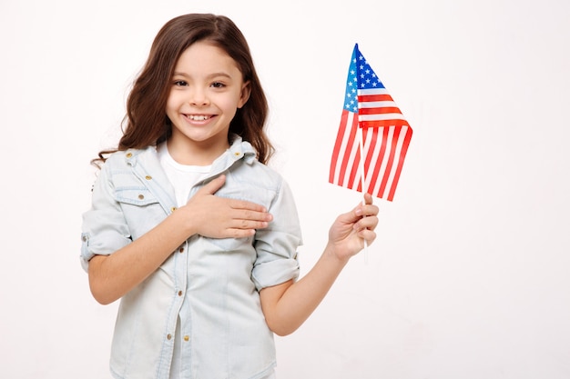 Vrolijk lachend gelukkig meisje dat de Amerikaanse vlag vasthoudt en haar borst aanraakt terwijl ze geluk uitdrukt en tegen een witte muur staat