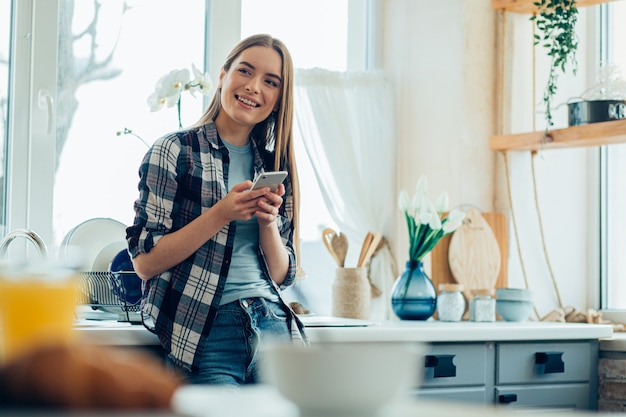 Vrolijk lachende jonge vrouw in de keuken staan met een smartphone. Websitebanner