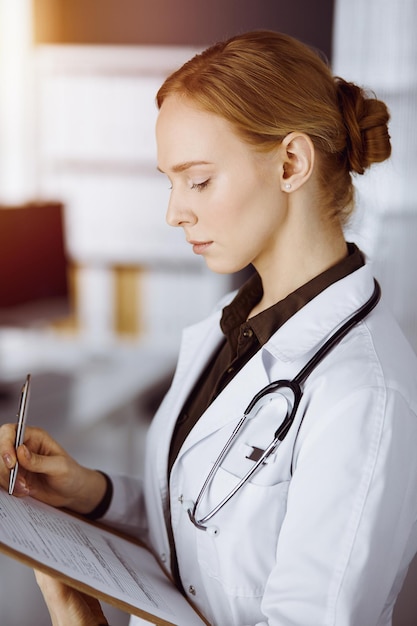 Vrolijk lachende vrouwelijke arts met klembord in zonnige kliniek. Portret van vriendelijke artsenvrouw op het werk. Geneeskunde concept.