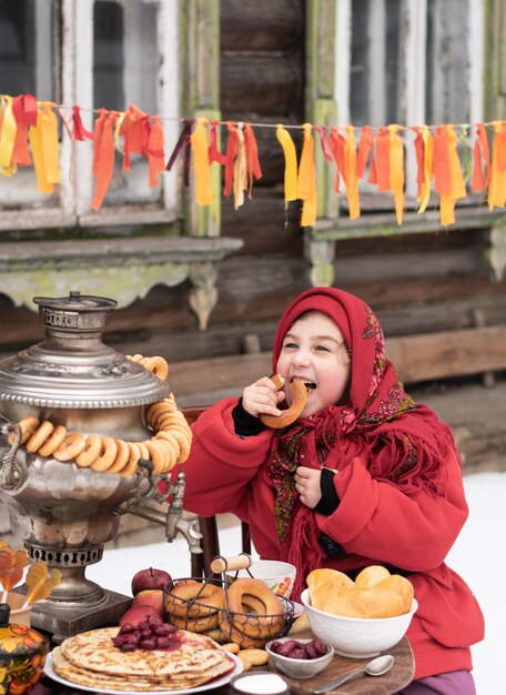 Vrolijk meisje bijt een grote bagel zit aan een tafel met een samovar en pannenkoeken