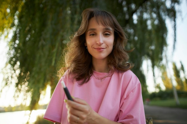 Vrolijk meisje in een roze t-shirt glimlach, met een elektronische sigaret, in het park.