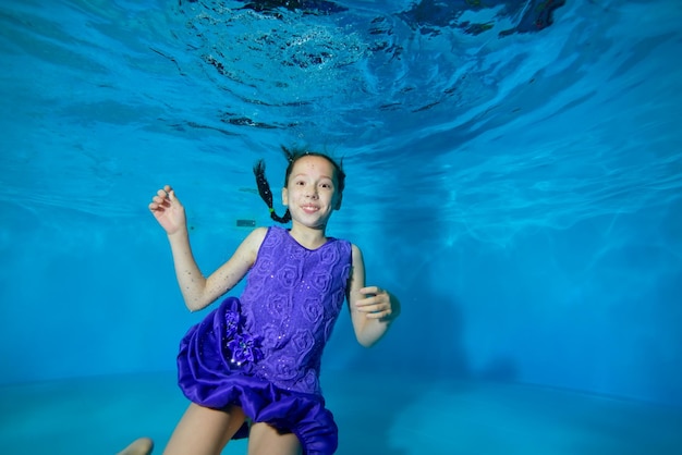 Vrolijk meisje in paarse jurk zwemt onder water in het zwembad op een blauwe achtergrond