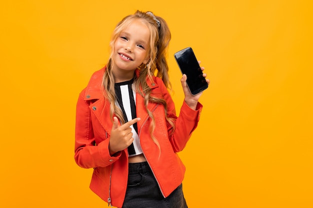 Vrolijk meisje in t-shirt en broek glimlacht met haar smartphone geïsoleerd op geel en oranje