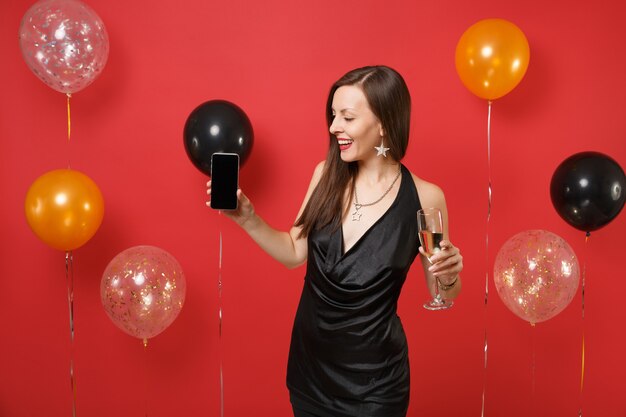 Vrolijk meisje in zwarte jurk houdt glas champagne vast, kijkend op mobiele telefoon met leeg zwart leeg scherm op heldere rode achtergrondluchtballonnen. Gelukkig Nieuwjaar, verjaardag mockup vakantie feest concept.